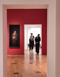 Photographer Peter Denton's image from the National Portrait Gallery, London's, exhibition, "Catherine Opie: To Be Seen." The image shows Catherine Opie's photo of marathon swimmer Diana Nyad, unclothed, seen from behind. It hangs on a red wall about 10 feet away. Nyad displays distinct tan lines on her back and backside. Two gallery visitors stand in a hallway to the right of Opie's photo. The exhibition closed on May 31, 2026.